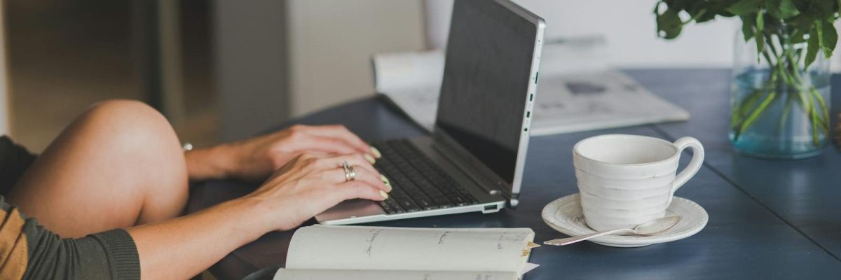 Female freelancer using laptop with coffee at home office desk, surrounded by roses and a planner.